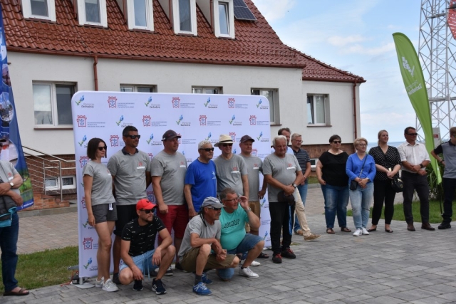 Zdjęcie grubowe, mężczyźni i kobieta będacy uczestnikami etapowych regat na dle ścianki z logiem Zachodniopomorskiego Okręgowego Związku Żeglarskiego wraz z logiem Pomorza Zachodniego.