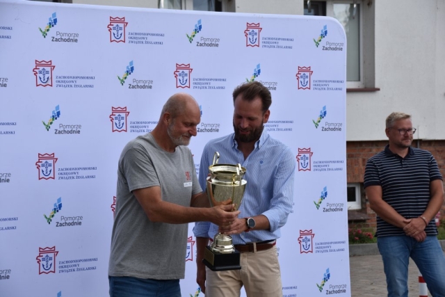 Wręczenie uczestnikowi 59. Etapowych Regat Turystycznych złotego pucharu na tle ścianki z logiem Zachodniopomorskiego Okręgowego Związku Żeglarskiego wraz z logiem Pomorza Zachodniego.