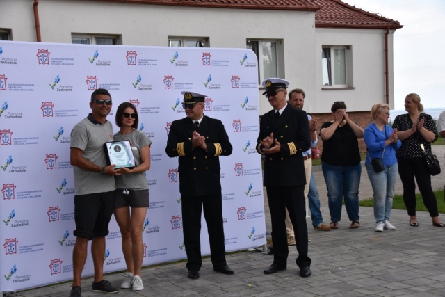 Para uczestników na tle ścianki z logiem Zachodniopomorskiego Okręgowego Związku Żeglarskiego wraz z logiem Pomorza Zachodniego pozuje do zdjęcia z dyplomem wręczonym od Marynarzy w ciemnych strojach, ze złotymi akcentami.