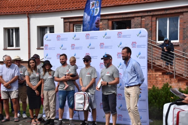 Członkowie 59. Etapowych Regat Turystycznych pozujący do grupowego zdjęcia na tle ścianki z logiem Zachodniopomorskiego Okręgowego Związku Żeglarskiego wraz z logiem Pomorza Zachodniego..