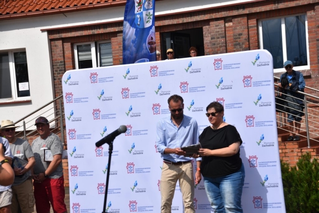 Kobieta będąca uczestniczką 59. Etapowych Regat Turystycznych pozująca z uzyskanym dyplomem na tle ścianki z logiem Zachodniopomorskiego Okręgowego Związku Żeglarskiego wraz z logiem Pomorza Zachodniego.