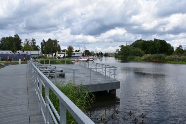 Rozlewisko rzeki Łarpia, po lewej stronie pomost spacerowy i taras widokowy, w tle przycumowane do przystani żaglówki i korony drzew oraz trzciny