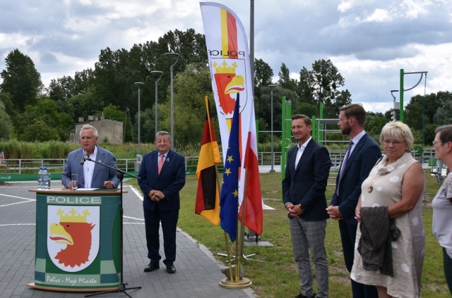 W plenerze, na chodniku po lewej stronie przy mównicy z grafiką gminy Police stoi mężczyzna w jasnym garniturze, przemawiający do mikrofonu, obok stoi drugi mężczyzna w ciemnym garniturze, stojak z flagami Niemiec, Unii Europejskiej i Polski, po prawej stronie stoją dwaj mężczyźni w ciemnych garniturach i kobieta w jasnej sukience, w tle siłownia pod chmurką i drzewa