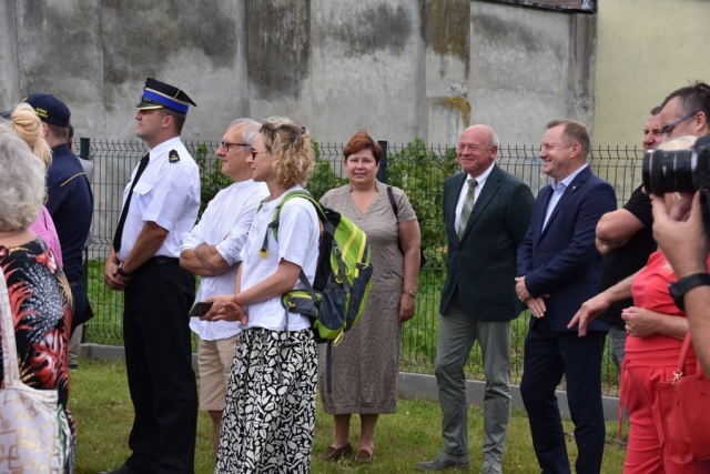 Na trawniku stoją zgromadzeni goście, mężczyzna w stroju galowym strażackim, dwaj mężczyźni w ciemnych garniturach, kobieta w jasnej sukience, kobieta w białej koszulce z plecakiem na plecach