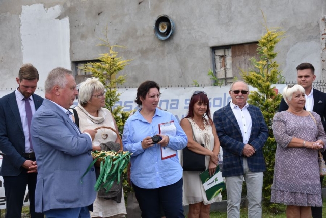 W jasnym garniturze stoi mężczyzna trzymający wiklinowy koszyczek z okolicznościowymi nożyczkami do przecięcia wstęgi, w tle stoją zaproszeni goście