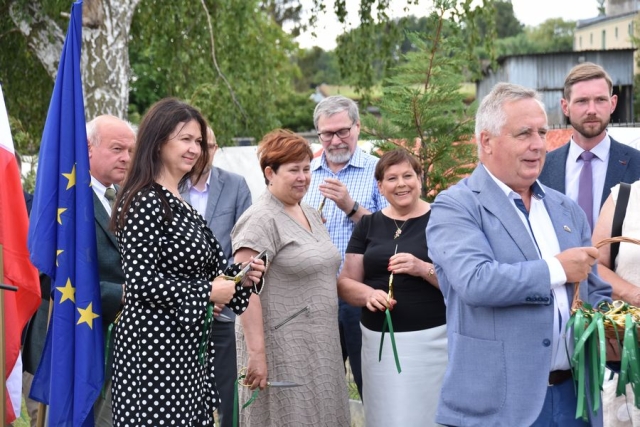 Przy fladze Unii Europejskiej, stoją zaproszeni goście, czekający do uroczystego przecięcia wstęgi, w dłoniach trzymają specjalnie przygotowane nożyczki