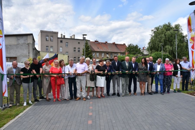 Na deptaku, przy rozpiętej od lewej do prawej szerokiej zielonej wstęgi, stoją zaproszeni goście trzymający w dłoniach nożyczki