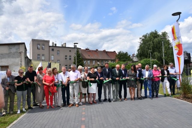 Na deptaku, stoją zaproszeni goście trzymający w dłoniach nożyczki w chwili przecinania rozpiętej na cała szerokość zielonej, szerokiej wstęgi