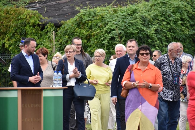 Na tle zielonej roslinności, zaproszeni goście udający się na spacer po nowo otwartej zakończonej inwestycji rewitalizacji terenów rekreacyjnych wzdłuż rzeki Łarpia