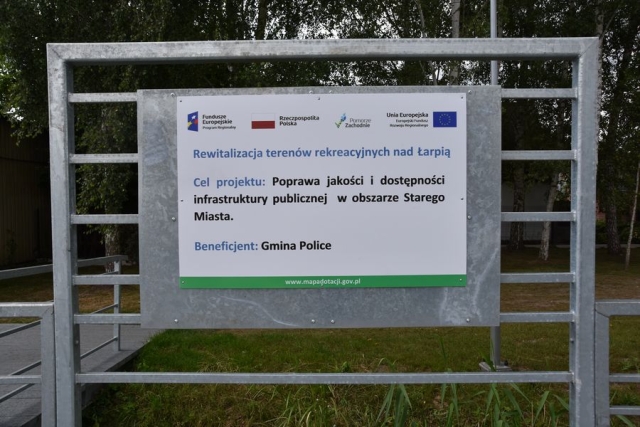 Biała tablica przymocowana do blachy metalowej na poręczy pomostu spacerowego wzdłuż rzeki, z informacją o projekcie Rewitalizacja terenów rekreacyjnych nad Łarpią, w tle zielona trawa i drzewa
