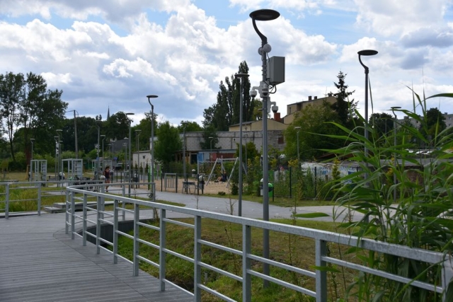 Pomost spacerowy przy rzece Łarpia, w tle plac zabaw i zabudowania oraz trzciny, drzewa