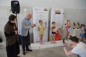 3 osoby stojące na podium, po lewej stronie 2 osoby bijące brawo