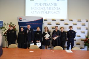 Stojące osoby w jednym rzędzie, w tle elementy reklamowe