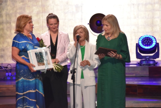 Cztery panie stoją na scenie, jedna z nich przemawia do stojącego mikrofonu