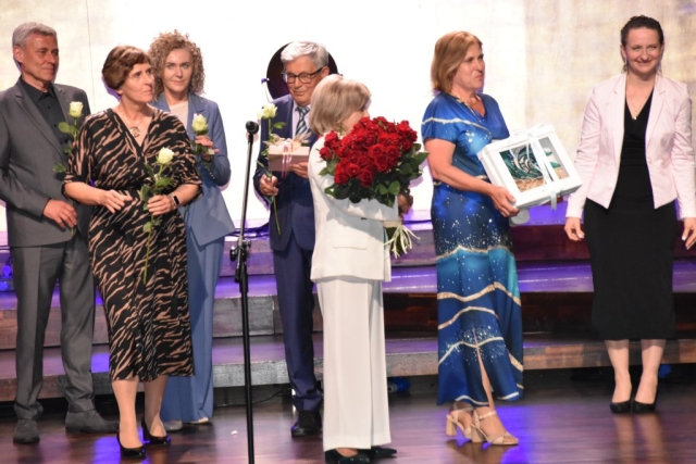 Wyróżnione osoby stoją na scenie, a przed nimi kobieta trzymająca duży bukiet czerwonych róż