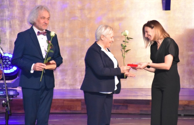 Kobieta wręcza pudełko z odznaczeniem drugiej kobiecie, obok stoi mężczyzna z różą w ręku