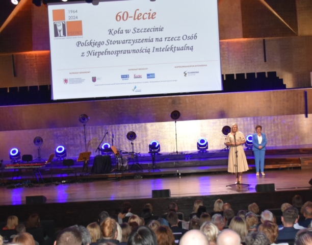 Na scenie stoją dwie panie, jedna z nich przemawia do stojącego mikrofonu