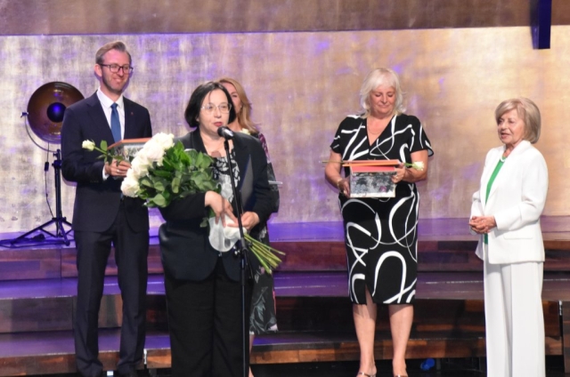 Kobieta przemawia do stojącego przed nią mikrofonu, za nią stoją trzy panie i jeden pan