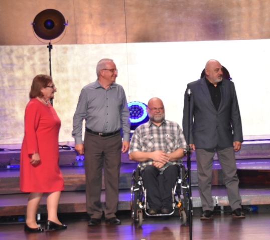 Na scenie stoją trzy osoby i jedna na wózku