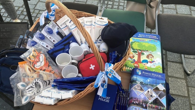 W koszu wiklinowym ułożone upominki Powiatu Polickiego w postaci między innymi kubeczków, opasek odblaskowych,, apteczek podecznych, kolorowanek