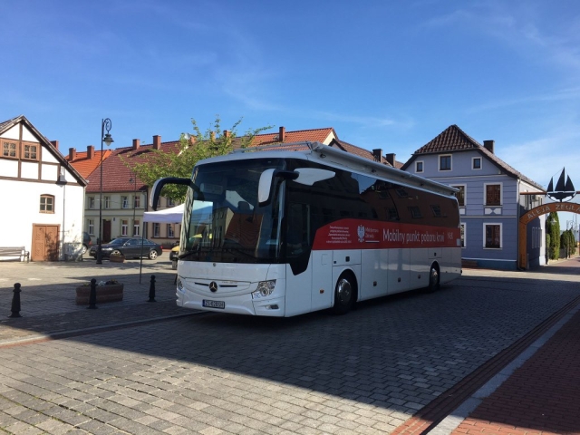 Autobus Mobilnego punktu poboru krwi stojący przy budynku, w tle zabudowania mieszkalne i zielone drzewo