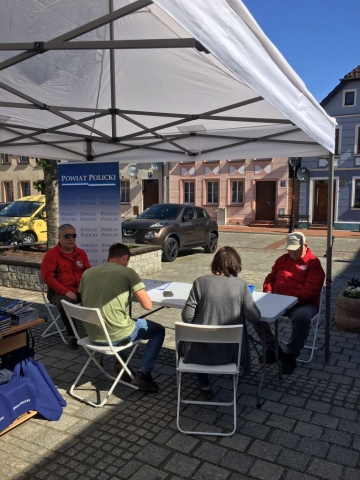 Osoby siedzące na krzesełkach przy stole, wypełniają formularze niezbędne do kwalifikacji by oddać krew, w tle zabudowania mieszkalne, a także zaparkowane dwa auta