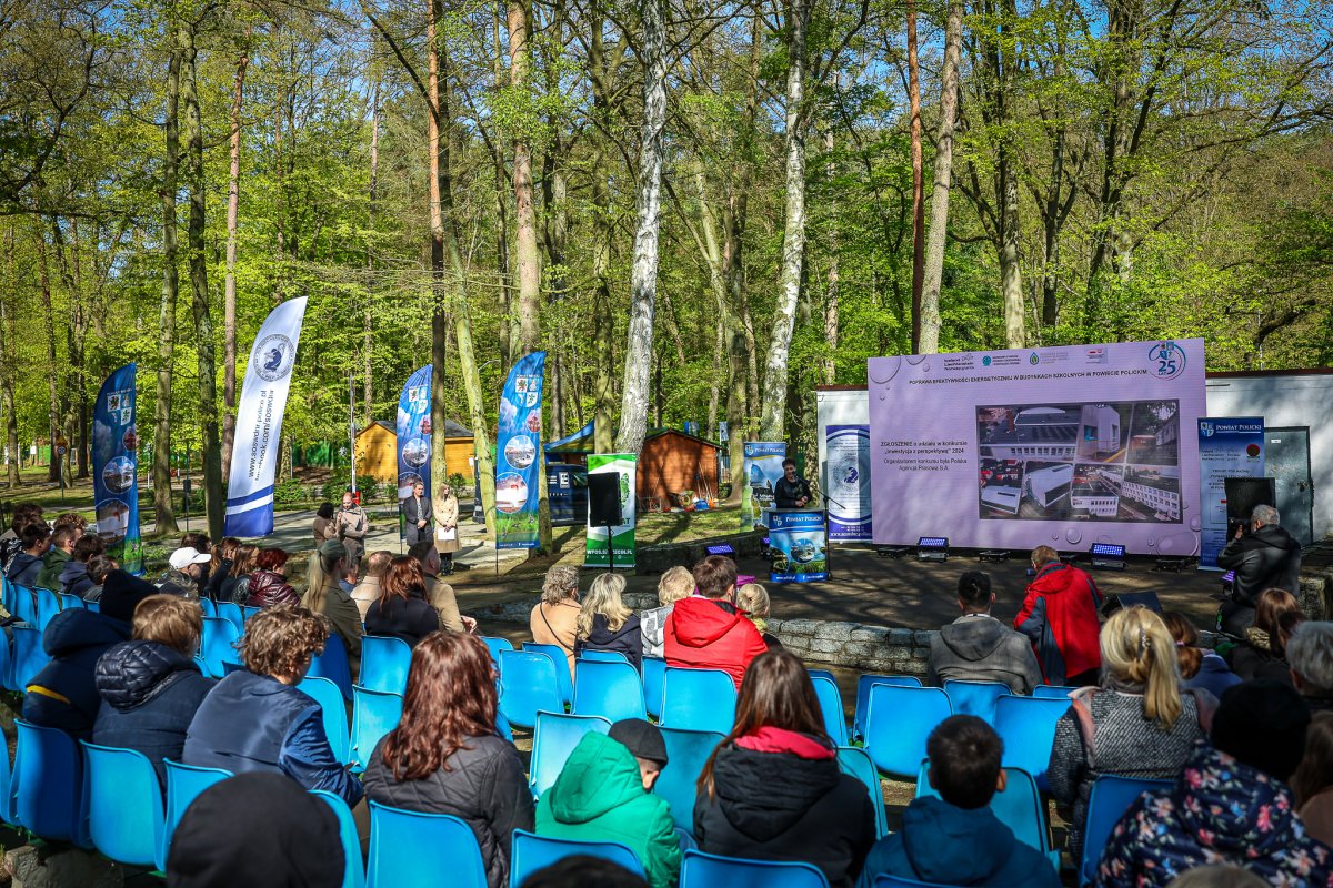 Na pierwszym planie siedzące osoby, w tle jedna osoba stojąca i telebim