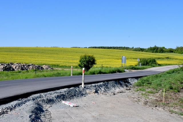 Remont drogi, trwają prace wiążące warstwę ścieralną jezdni, w tle pole rzepakowe zielone drzewa i krzewy