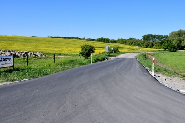 Remont drogi, trwają prace wiążące warstwę ścieralną jezdni, w tle pole rzepakowe zielone drzewa i krzewy.