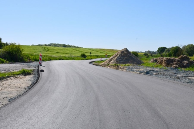 Remont drogi, trwają prace wiążące warstwę ścieralną jezdni, w tle hałdy żwiru i piasku oraz pole, zielone drzewa i krzewy