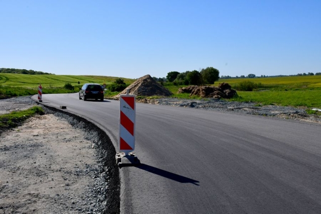 Remont drogi, trwają prace wiążące warstwę ścieralną jezdni, w tle hałdy żwiru i piasku oraz pole, zielone drzewa i krzewy