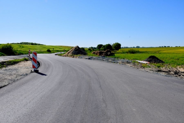 Remont drogi, trwają prace wiążące warstwę ścieralną jezdni, w tle hałdy żwiru i piasku oraz pole, zielone drzewa i krzewy