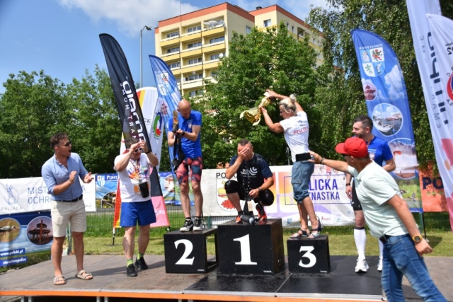 Uczestnik kucający na podium schładzany jest wodą z dużego pucharu przez organizatora