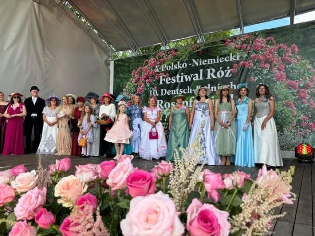 Na scenie stoją w kolorowych sukienkach kobiety, a także mężczyna w smokingu i kapeluszu