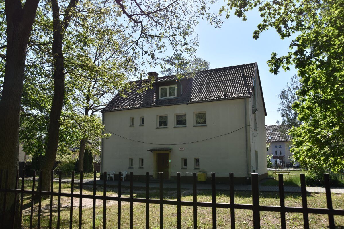 Dwukondygnacyjny budynek ze spadzistym dachem w otoczeniu zieleni, ogrodzony metalowym płotem.