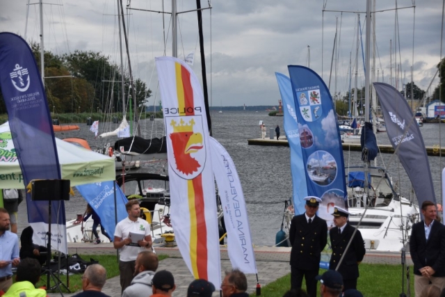 Windery na tle wody z żaglówkami, uczestnicy wydarzenia stoją na placu